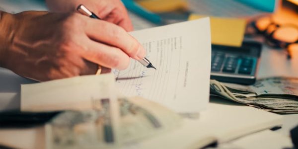 A hand signing financial documents on a desk with cash and a calculator, signifying finance management.